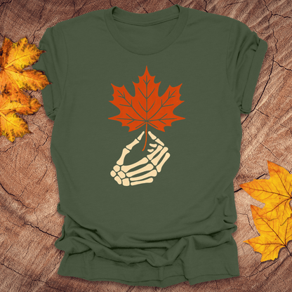 Green t-shirt with a skeleton hand holding an orange maple leaf on a wooden surface with fall leaves.