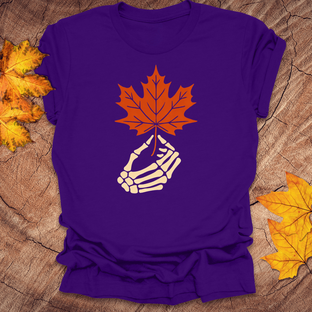 Purple t-shirt with a skeleton hand holding an orange leaf design on a wooden surface with fall leaves.