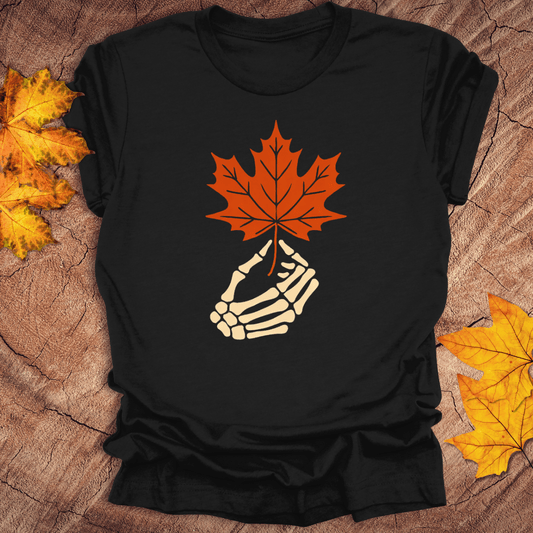 Black t-shirt with skeleton hand holding a red maple leaf on a wooden surface with fall leaves.