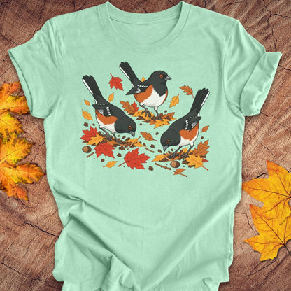 A mint green crewneck t-shirt featuring a graphic print of three towhees foraging among orange and red autumn leaves and acorns, laid flat on a rustic wood background with scattered maple leaves.