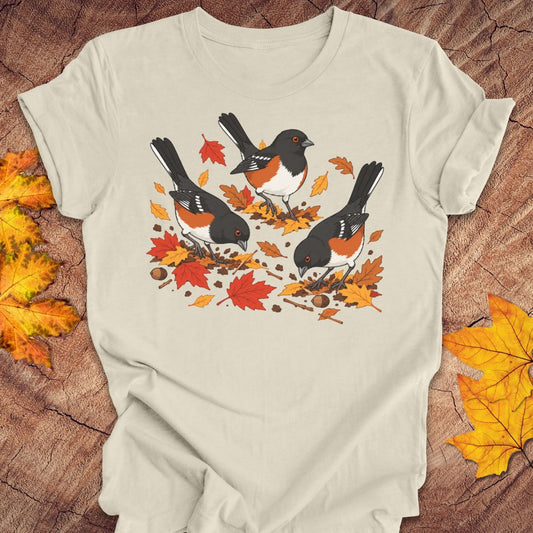 A light beige crewneck t-shirt featuring a graphic print of three towhees foraging among orange and red autumn leaves and acorns, laid flat on a rustic wood background with scattered maple leaves.
