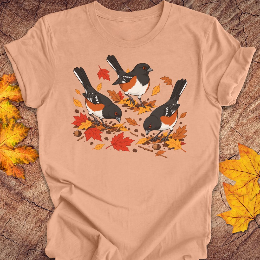 A peach crewneck t-shirt featuring a graphic print of three towhees foraging among orange and red autumn leaves and acorns, laid flat on a rustic wood background with scattered maple leaves.