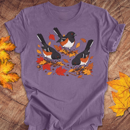 A purple crewneck t-shirt featuring a graphic print of three towhees foraging among orange and red autumn leaves and acorns, laid flat on a rustic wood background with scattered maple leaves.