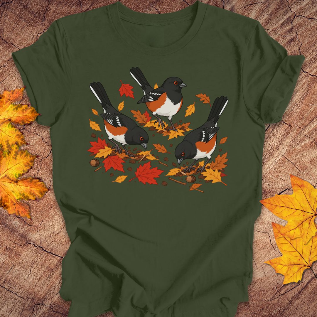 A green crewneck t-shirt featuring a graphic print of three towhees foraging among orange and red autumn leaves and acorns, laid flat on a rustic wood background with scattered maple leaves.