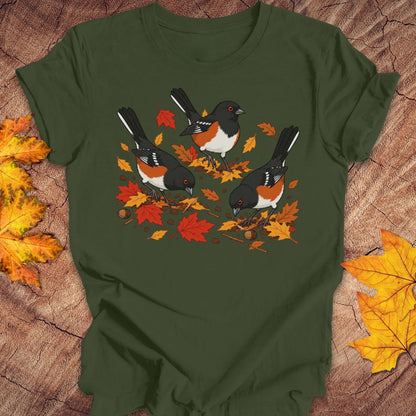A green crewneck t-shirt featuring a graphic print of three towhees foraging among orange and red autumn leaves and acorns, laid flat on a rustic wood background with scattered maple leaves.