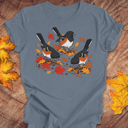 A blue crewneck t-shirt featuring a graphic print of three towhees foraging among orange and red autumn leaves and acorns, laid flat on a rustic wood background with scattered maple leaves.