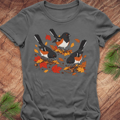 A dark gray short-sleeve t-shirt with a graphic of three towhee birds pecking at leaves on the ground, laid flat on a rustic wood background with pine branches.