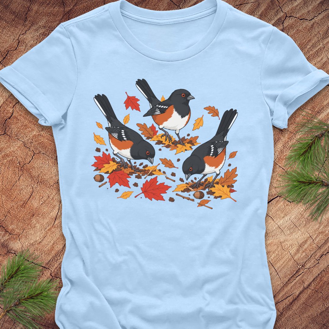 A dark gray short-sleeve t-shirt with a graphic of three towhee birds pecking at leaves on the ground, laid flat on a rustic wood background with pine branches.