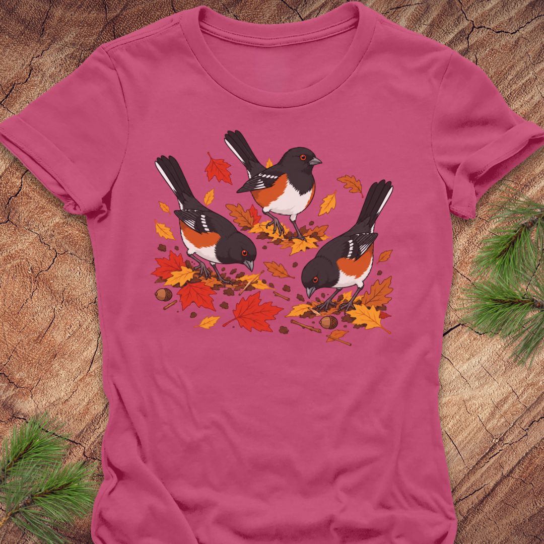 A pink short-sleeve t-shirt with a graphic of three towhee birds pecking at leaves on the ground, laid flat on a rustic wood background with pine branches.