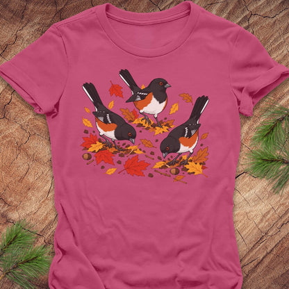 A pink short-sleeve t-shirt with a graphic of three towhee birds pecking at leaves on the ground, laid flat on a rustic wood background with pine branches.