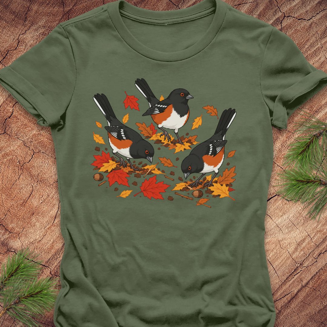 A green short-sleeve t-shirt with a graphic of three towhee birds pecking at leaves on the ground, laid flat on a rustic wood background with pine branches.