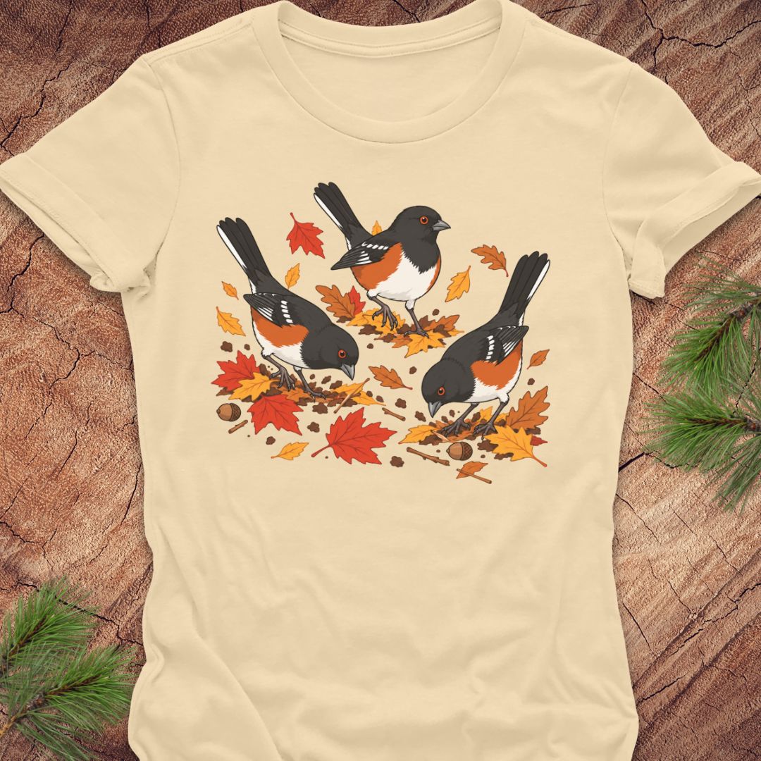 A cream colored short-sleeve t-shirt with a graphic of three towhee birds pecking at leaves on the ground, laid flat on a rustic wood background with pine branches.