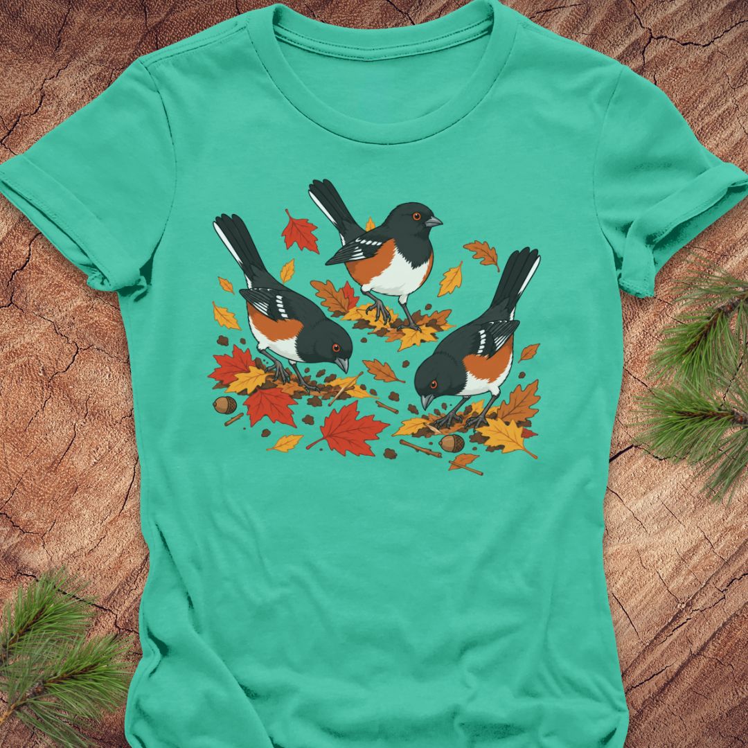 A mint green short-sleeve t-shirt with a graphic of three towhee birds pecking at leaves on the ground, laid flat on a rustic wood background with pine branches.