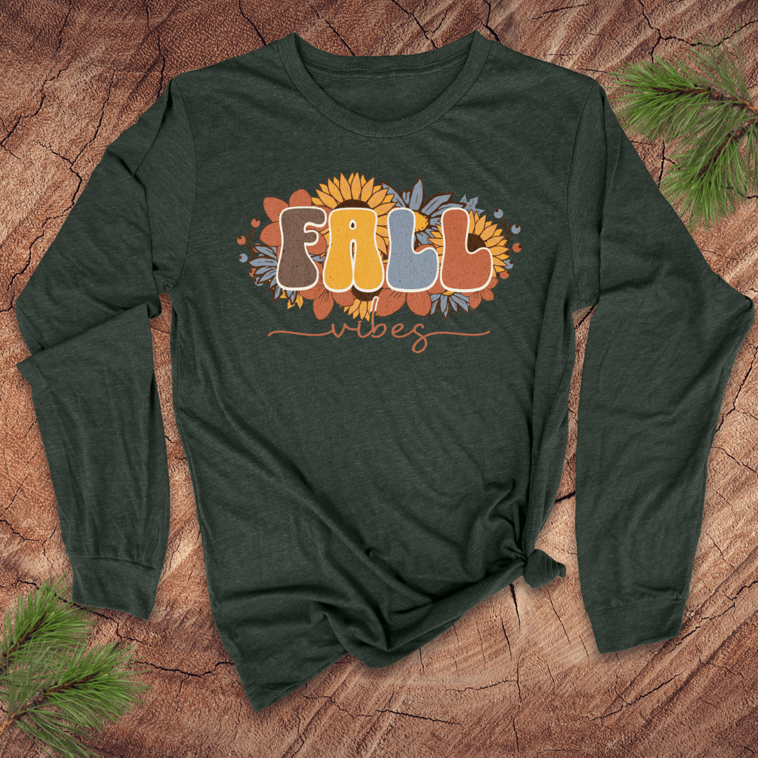 Green long-sleeve shirt with 'Fall Vibes' text on a wooden surface with pine branches.