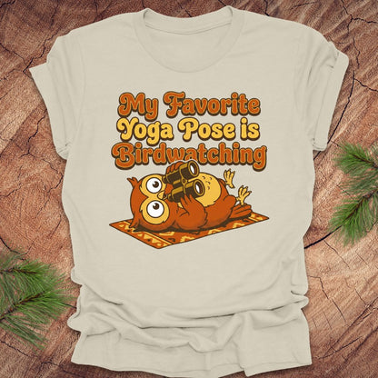 Beige t-shirt with owl design and text 'My Favorite Yoga Pose is Birdwatching' on a wooden surface.