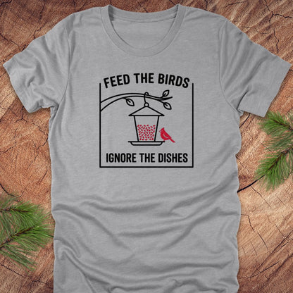 Gray t-shirt with a graphic design of a bird feeder and text 'feed the birds. ignore the dishes' on a wooden surface.
