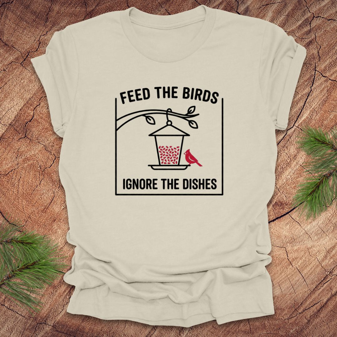Beige t-shirt with a graphic design of a bird feeder and text 'feed the birds. ignore the dishes' on a wooden surface.