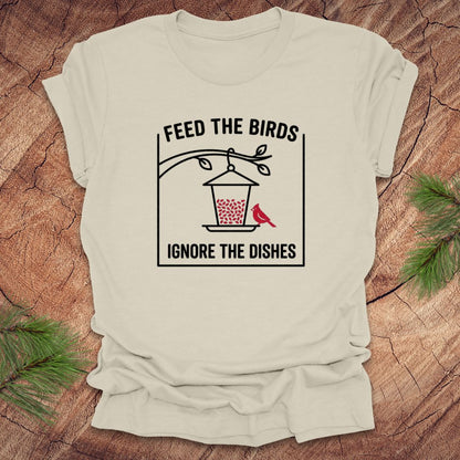 Beige t-shirt with a graphic design of a bird feeder and text 'feed the birds. ignore the dishes' on a wooden surface.