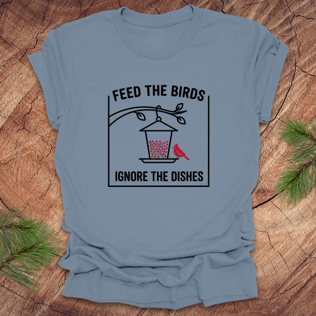Steel blue t-shirt with a graphic design of a bird feeder and text 'feed the birds. ignore the dishes' on a wooden surface.