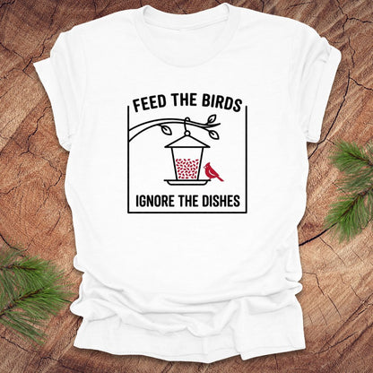 White  t-shirt with a graphic design of a bird feeder and text 'feed the birds. ignore the dishes' on a wooden surface.