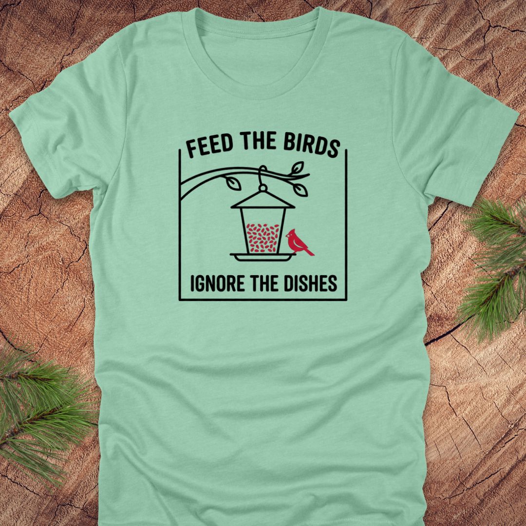 Mint green t-shirt with a graphic design of a bird feeder and text 'feed the birds. ignore the dishes' on a wooden surface.
