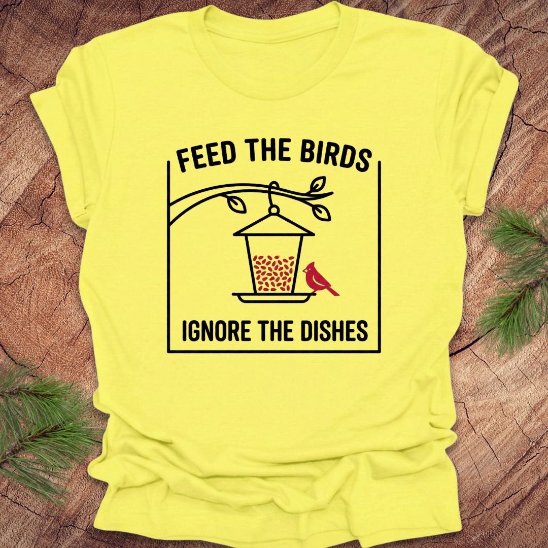 Yellow t-shirt with a graphic and text 'Feed the birds ignore the dishes' on a wooden surface.