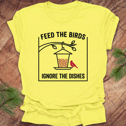 Yellow t-shirt with a graphic and text 'Feed the birds ignore the dishes' on a wooden surface.