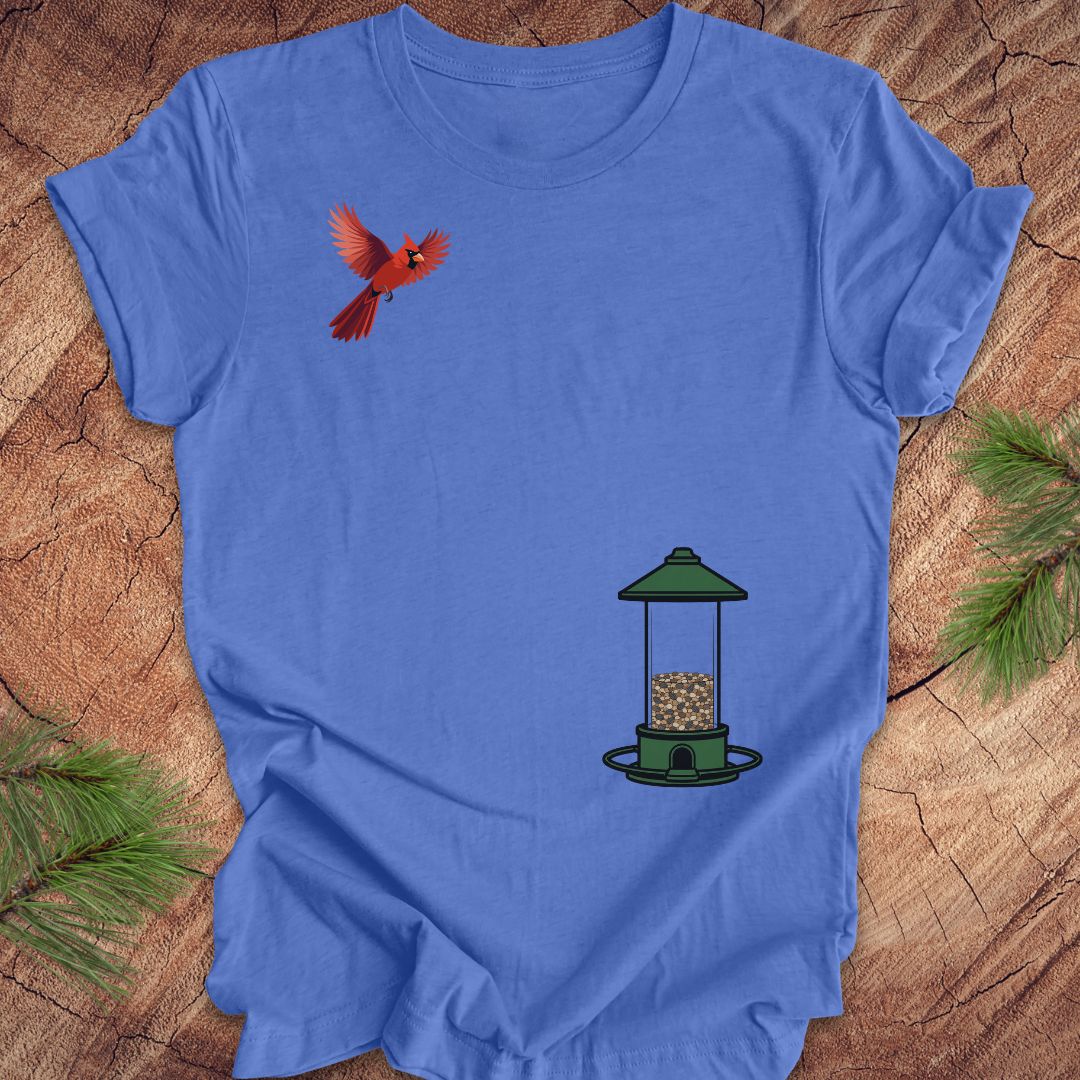 A blue short-sleeve crewneck t-shirt laid flat on a rustic wood background with pine branches in the corners. The shirt features a graphic print of a red cardinal bird flying near the upper left chest and a green bird feeder with seed near the lower right chest.