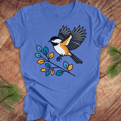 A blue short-sleeve crewneck t-shirt featuring a graphic of a chickadee bird perched on a branch with colorful leaves, laid flat on a rustic wood background with pine needles in the corners.