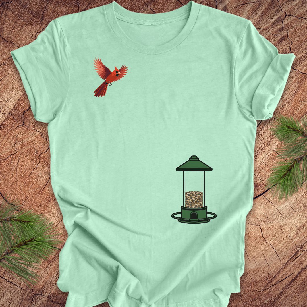 A mint green short-sleeve t-shirt laid flat on a rustic wood surface, featuring a red cardinal bird graphic on the upper left chest and a green bird feeder graphic on the lower right chest.