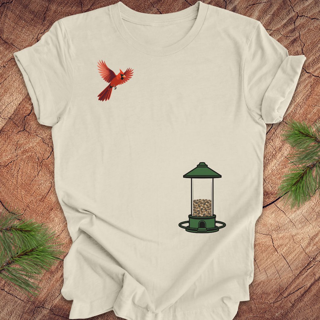 A beige short-sleeve t-shirt laid flat on a rustic wood background, featuring a graphic of a red cardinal flying on the upper left chest and a green bird feeder with seed on the lower right chest.