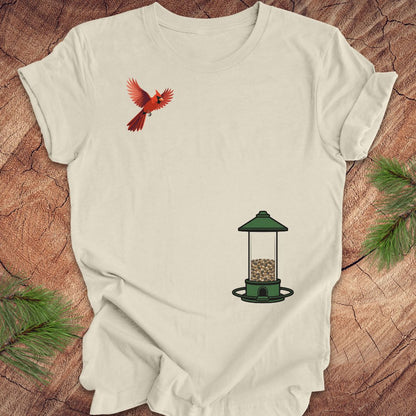 A beige short-sleeve t-shirt laid flat on a rustic wood background, featuring a graphic of a red cardinal flying on the upper left chest and a green bird feeder with seed on the lower right chest.