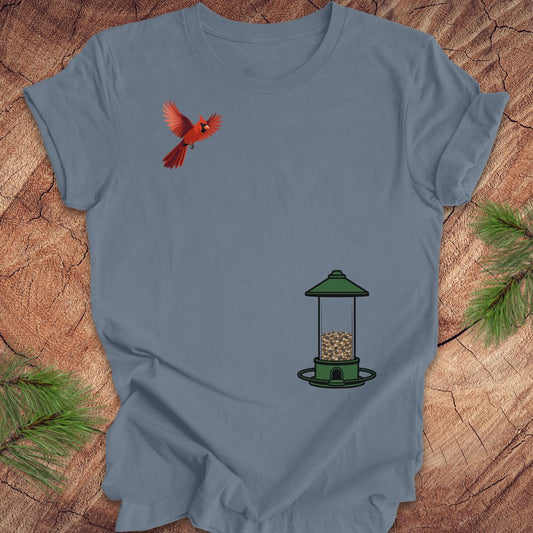 A slate blue short-sleeve t-shirt laid flat on a rustic wood background with pine branches in the corners. The shirt features a graphic of a red cardinal bird flying above a green bird feeder.