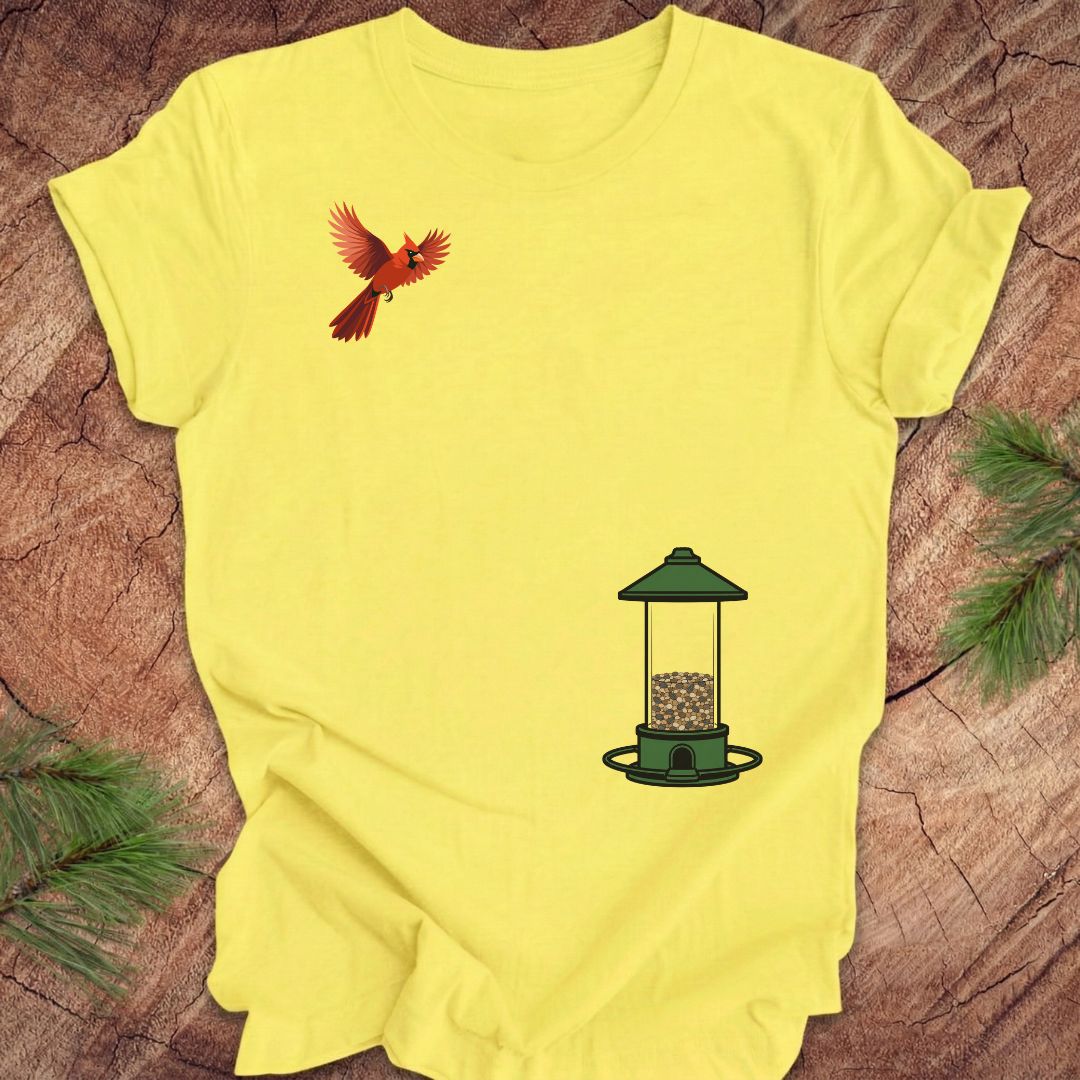 A bright yellow short-sleeve t-shirt laid flat on a textured wood surface, featuring a graphic of a red cardinal bird on the left chest and a green bird feeder on the lower right. Pine branches are visible in the corners.