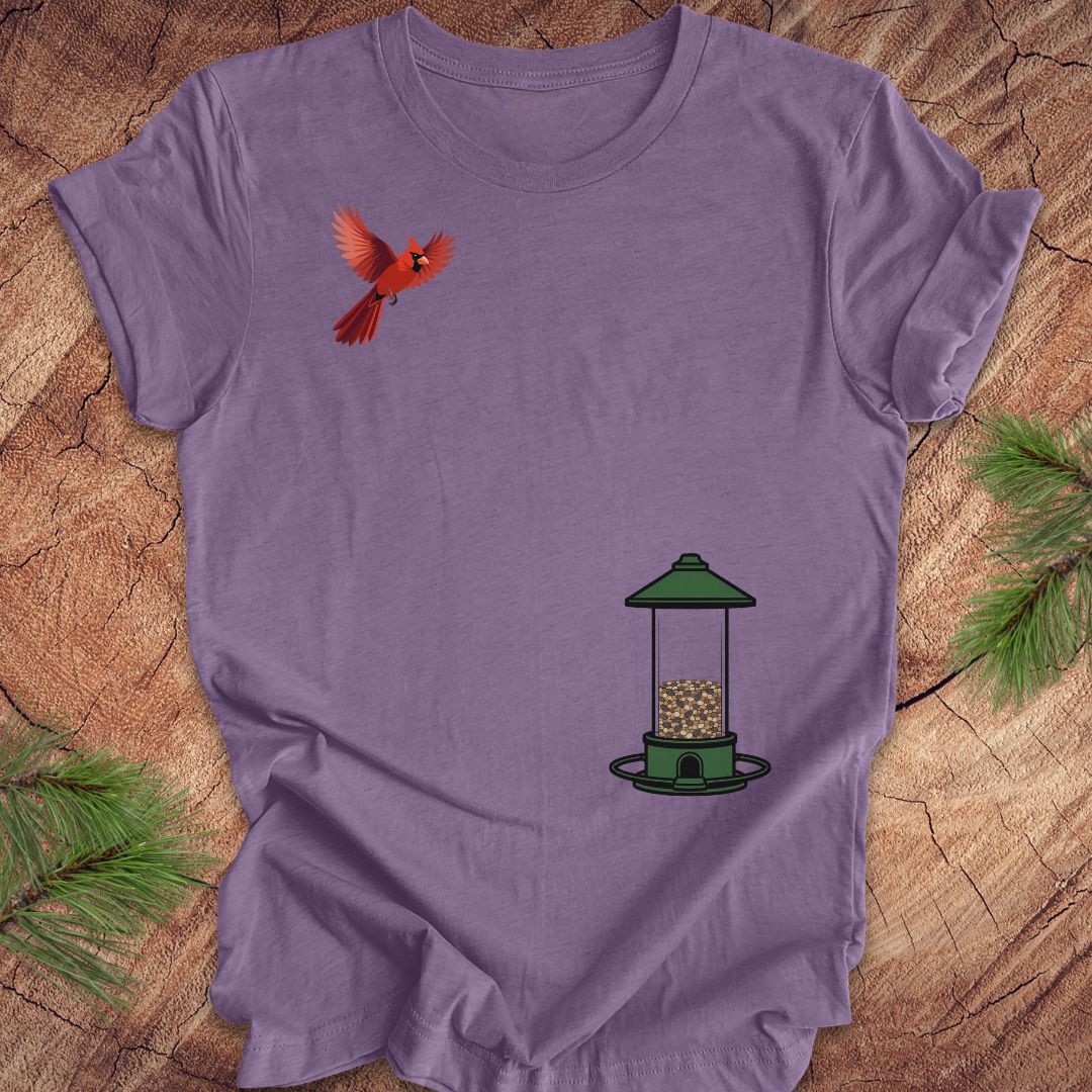 A lavender purple short-sleeve t-shirt laid flat on a rustic wood background with pine branches in the corners. The shirt features a graphic print of a red cardinal on the upper left chest and a green bird feeder with seeds on the lower right chest.