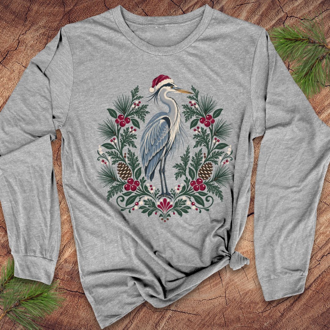 Athletic heather gray long-sleeve t-shirt with a Great Blue Heron in a Santa hat inside a folk-art wreath of pine, red berries, and pinecones, laid flat on a rustic wood background with pine branches.