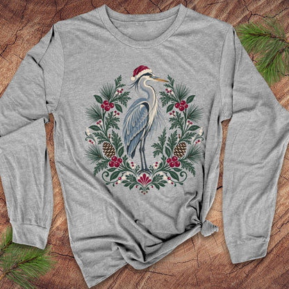 Athletic heather gray long-sleeve t-shirt with a Great Blue Heron in a Santa hat inside a folk-art wreath of pine, red berries, and pinecones, laid flat on a rustic wood background with pine branches.