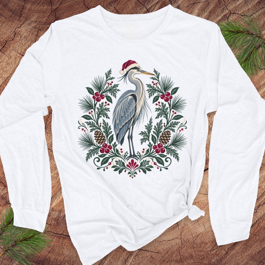 White long-sleeve t-shirt with a Great Blue Heron in a Santa hat inside a folk-art wreath of pine, red berries, and pinecones, laid flat on a rustic wood background with pine branches.