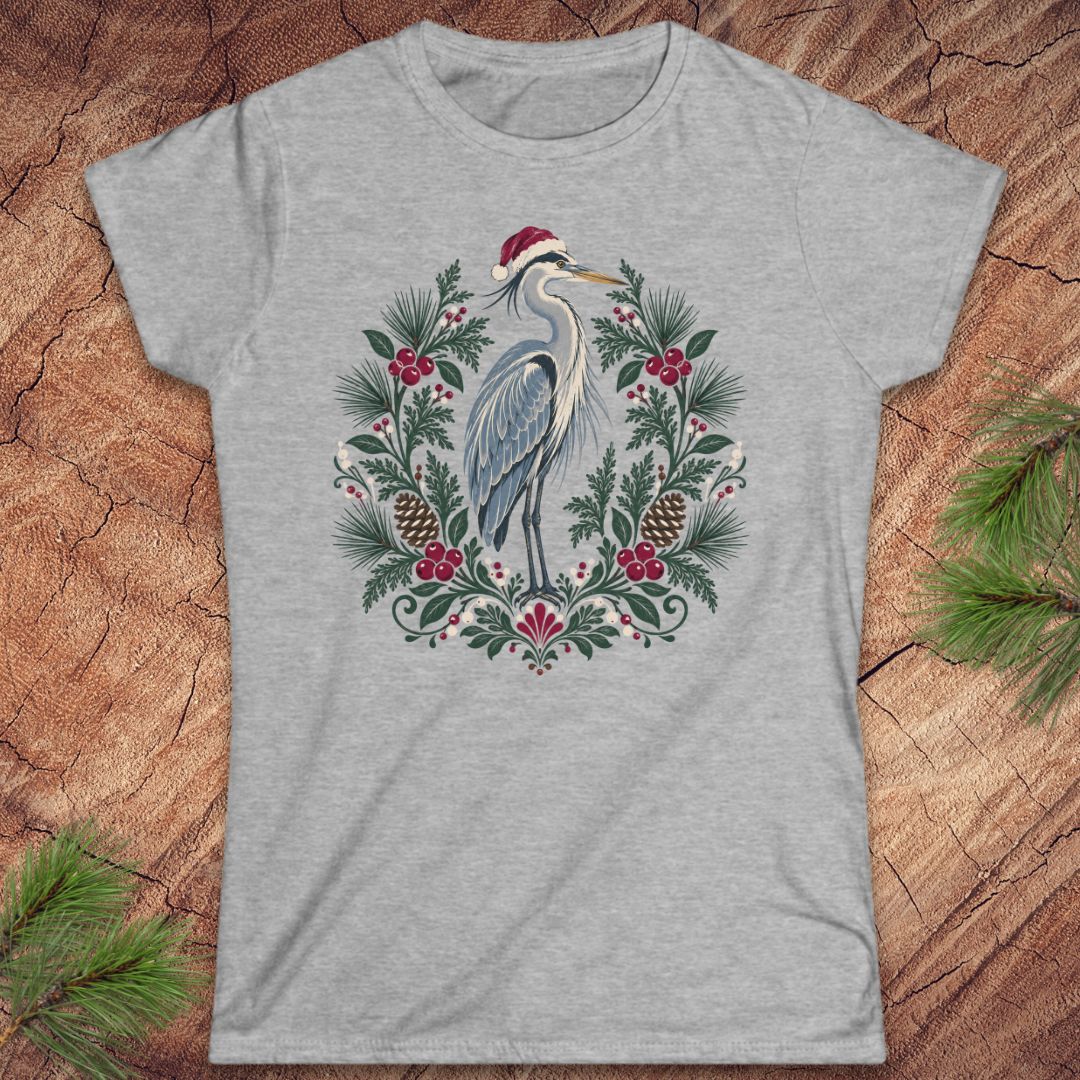 Sport grey women's t-shirt with a Great Blue Heron in a Santa hat inside a folk-art wreath of pine, red berries, and pinecones, laid flat on a rustic wood background with pine branches.