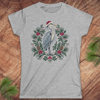 Sport grey women's t-shirt with a Great Blue Heron in a Santa hat inside a folk-art wreath of pine, red berries, and pinecones, laid flat on a rustic wood background with pine branches.