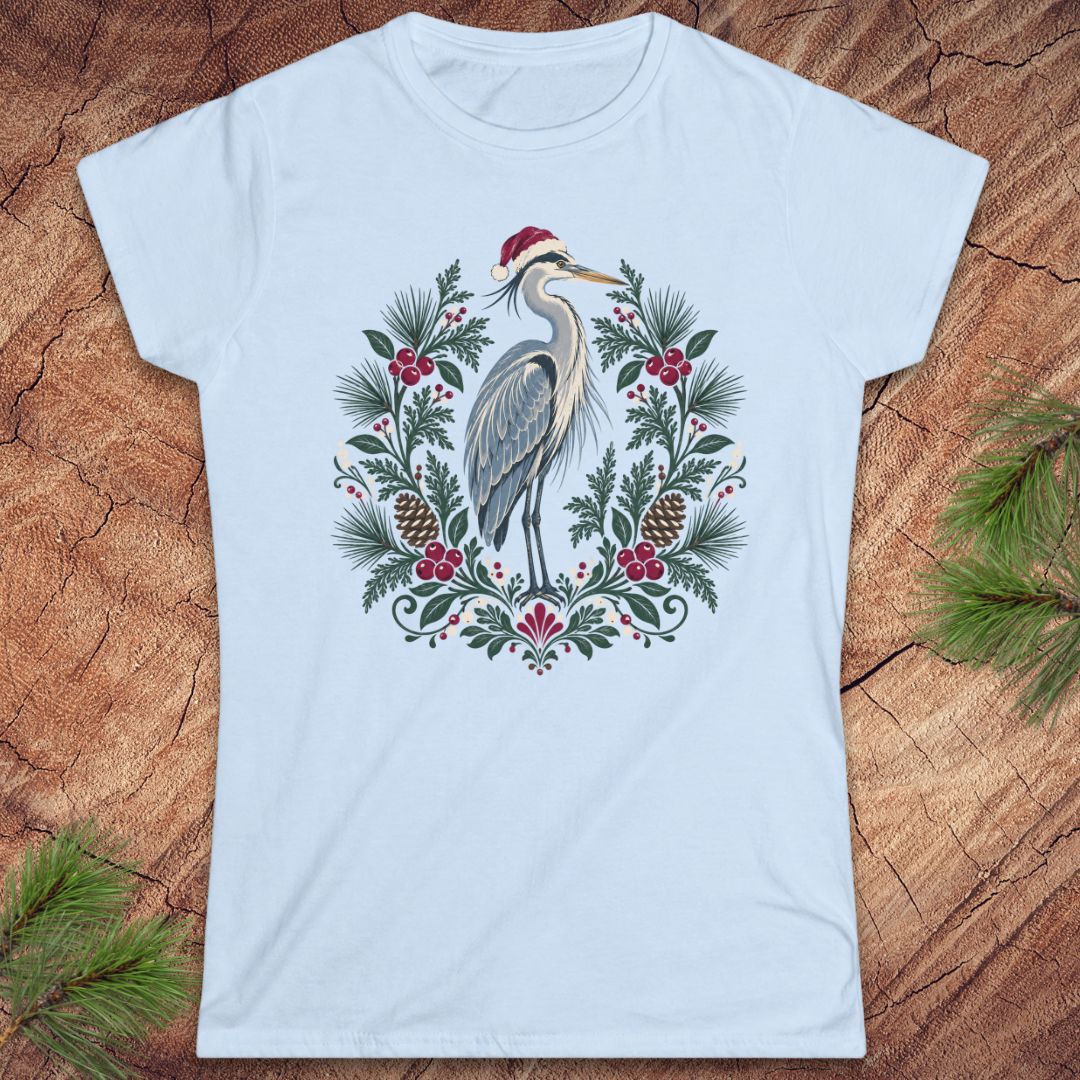 Light blue women's t-shirt with a Great Blue Heron in a Santa hat inside a folk-art wreath of pine, red berries, and pinecones, laid flat on a rustic wood background with pine branches.