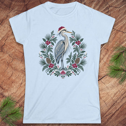 Light blue women's t-shirt with a Great Blue Heron in a Santa hat inside a folk-art wreath of pine, red berries, and pinecones, laid flat on a rustic wood background with pine branches.