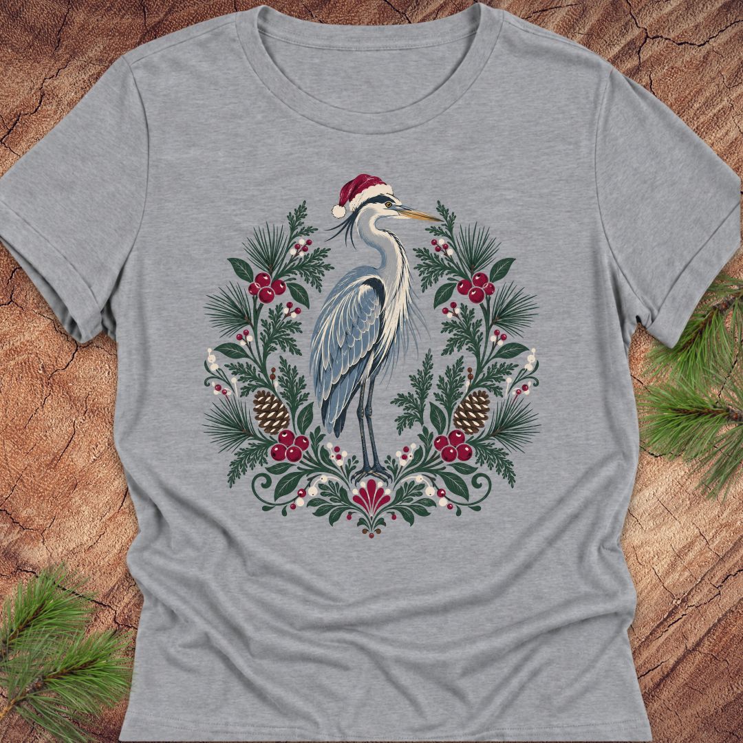 A heather gray short-sleeve t-shirt featuring a detailed graphic of a blue heron wearing a red Santa hat, surrounded by a festive wreath of green pine needles, red berries, and pinecones, laid flat on a rustic wood background with pine branches in the corners.