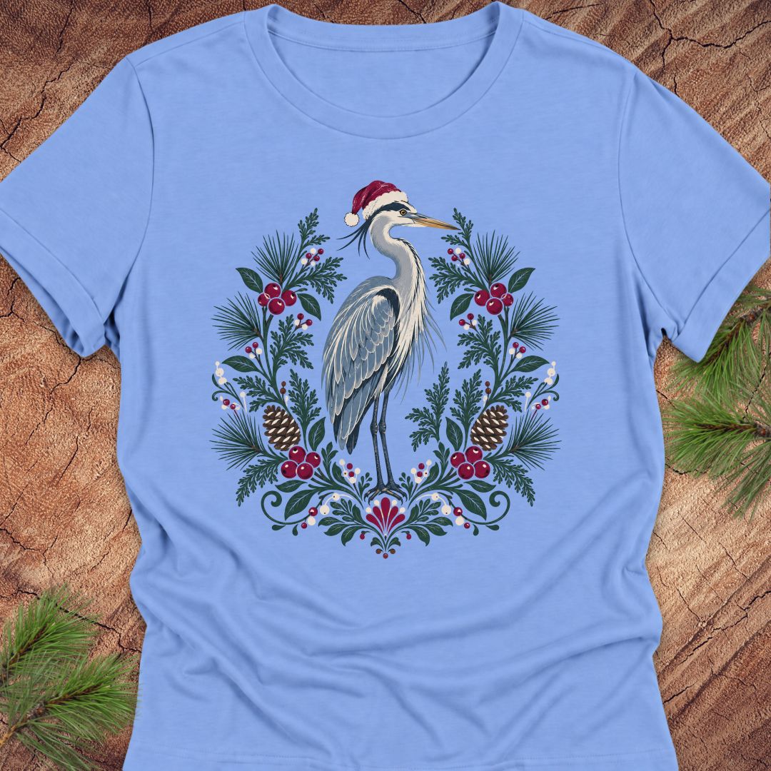 A light blue short-sleeve crewneck t-shirt featuring a festive graphic of a heron wearing a Santa hat, surrounded by a Christmas wreath with pinecones, berries, and pine needles, laid flat on a rustic wood background with pine branches.