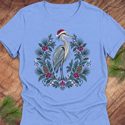 A light blue short-sleeve crewneck t-shirt featuring a festive graphic of a heron wearing a Santa hat, surrounded by a Christmas wreath with pinecones, berries, and pine needles, laid flat on a rustic wood background with pine branches.