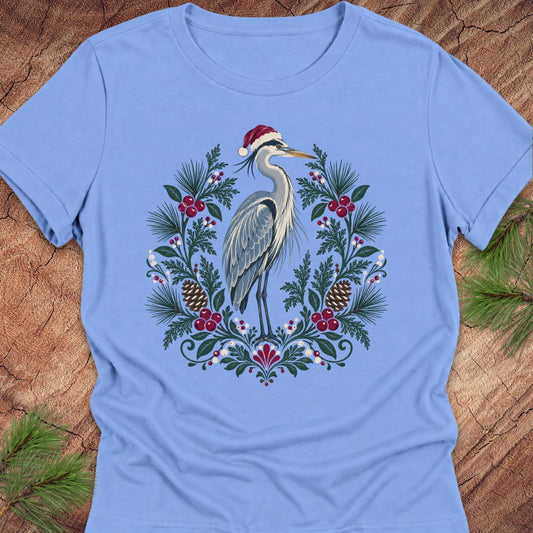 A light blue short-sleeve crewneck t-shirt featuring a festive graphic of a heron wearing a Santa hat, surrounded by a Christmas wreath with pinecones, berries, and pine needles, laid flat on a rustic wood background with pine branches.