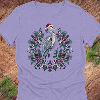 A lavender purple short-sleeve crewneck t-shirt featuring a festive graphic of a heron wearing a Santa hat, surrounded by a wreath of pine branches, pinecones, and red berries, laid flat on a rustic wood background with pine needles in the corners.