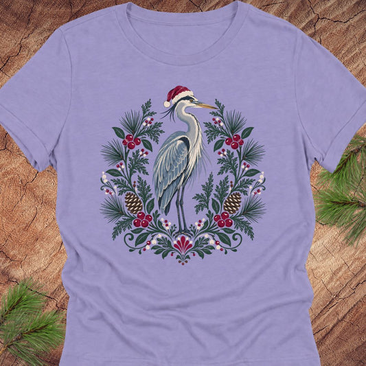 A lavender purple short-sleeve crewneck t-shirt featuring a festive graphic of a heron wearing a Santa hat, surrounded by a wreath of pine branches, pinecones, and red berries, laid flat on a rustic wood background with pine needles in the corners.