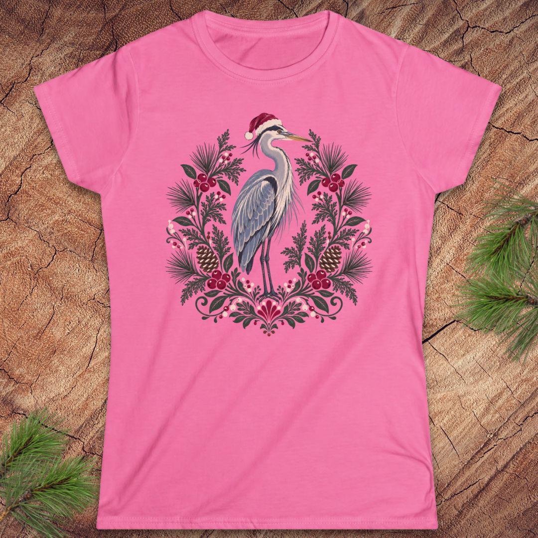 Azalea pink women's t-shirt with a Great Blue Heron in a Santa hat inside a folk-art wreath of pine, red berries, and pinecones, laid flat on a rustic wood background with pine branches.
