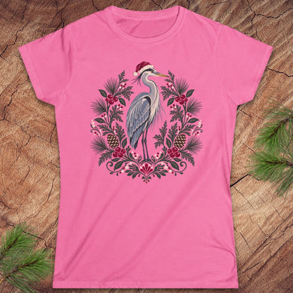 Azalea pink women's t-shirt with a Great Blue Heron in a Santa hat inside a folk-art wreath of pine, red berries, and pinecones, laid flat on a rustic wood background with pine branches.